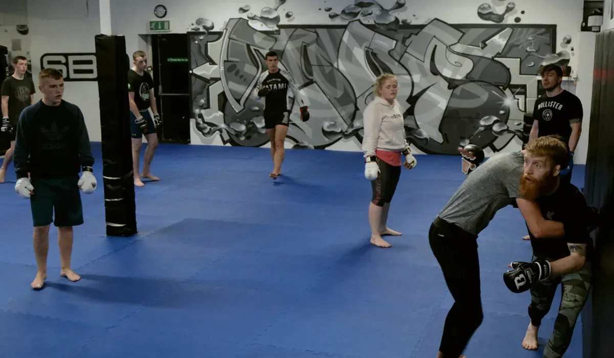 Paddy Holohan showing a clinch in the cage while the class watches on the blue mats